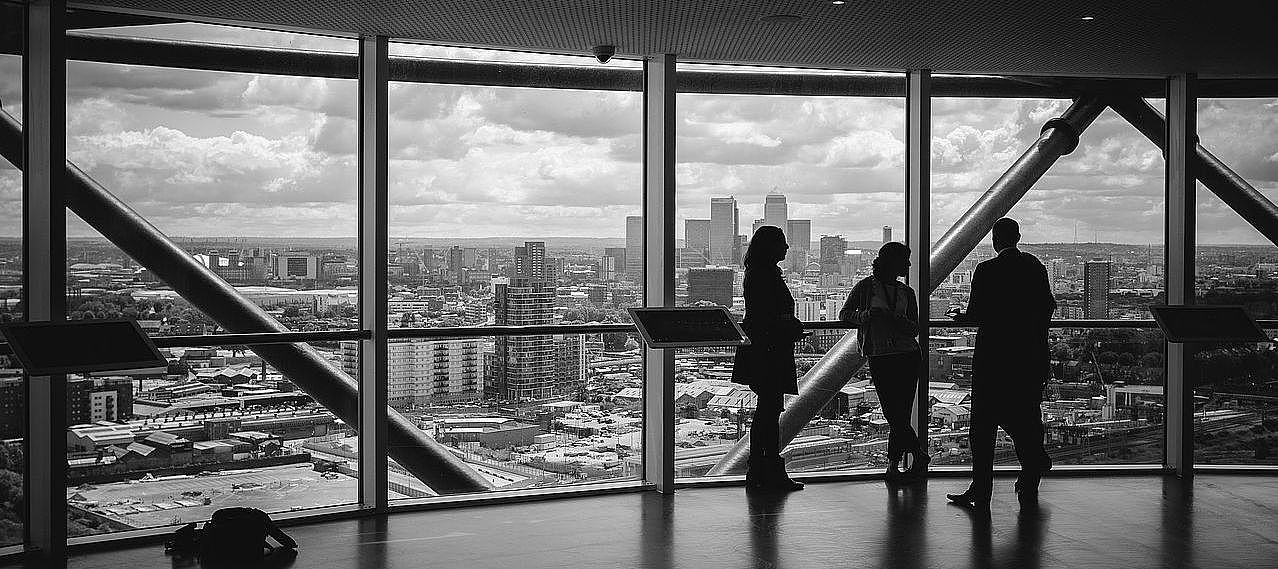 Schwarz-Weiß-Bild: Drei Personen unterhalten sich in einem Hochhaus vor einer Glasfassade, im Hintergrund ist die Skyline einer Großstadt zu sehen